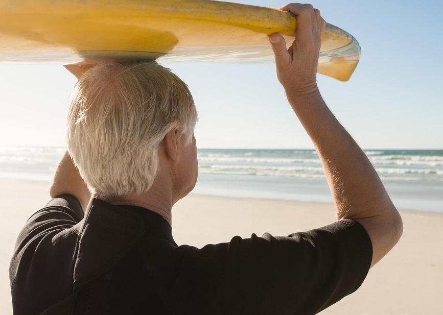 Rear view of senior man carrying surfboard on head at beach during sunny day