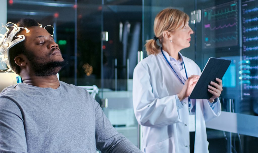 Man Wearing Brainwave Scanning Headset Sits in a Chair while Scientist with Tablet Computer Supervises Process. In the Modern Brain Study Laboratory Monitors Show EEG Reading and Brain Model.